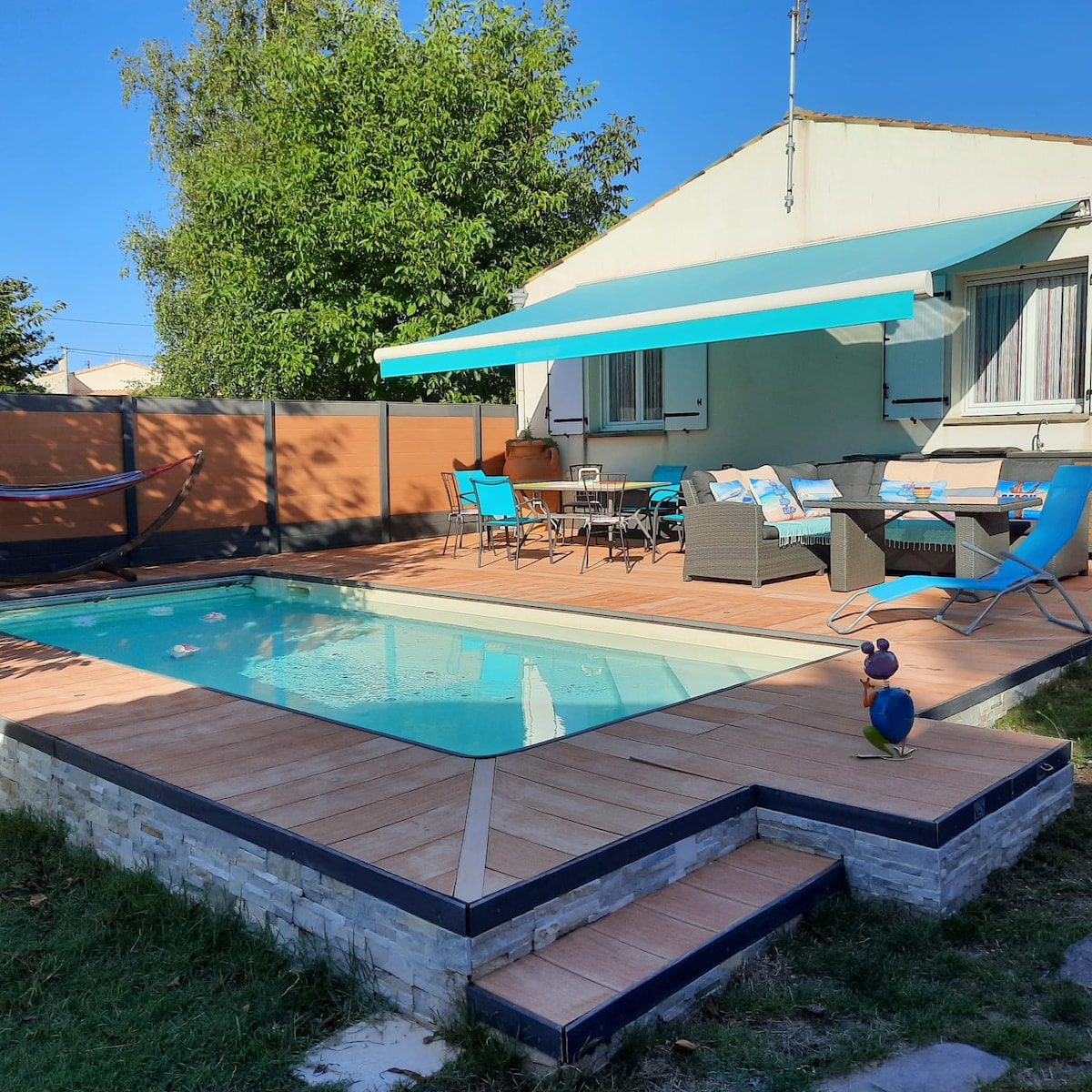 A private pool is surrounded by wooden decking and partial walls, offering a peaceful atmosphere. Comfortable outdoor seating and a dining area are shaded by a blue awning. A hammock is visible in the background, inviting relaxation. A small decorative figure adds a touch of charm.