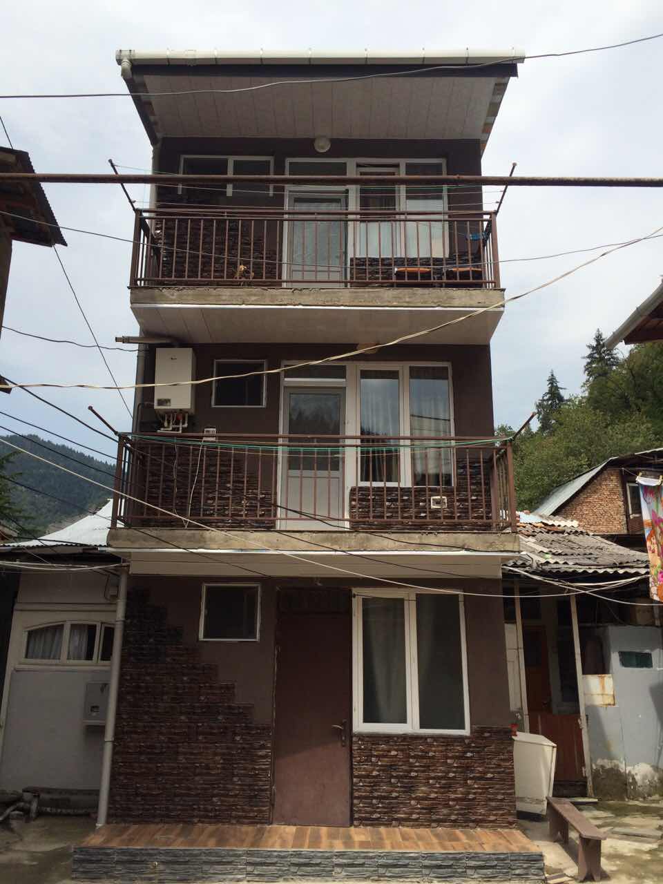A three-story building featuring a combination of wood and stone materials. Each floor is adorned with a set of glass windows and balcony railings. A main entrance door is situated at the base, with the surrounding area displaying a mix of rustic structures and greenery.