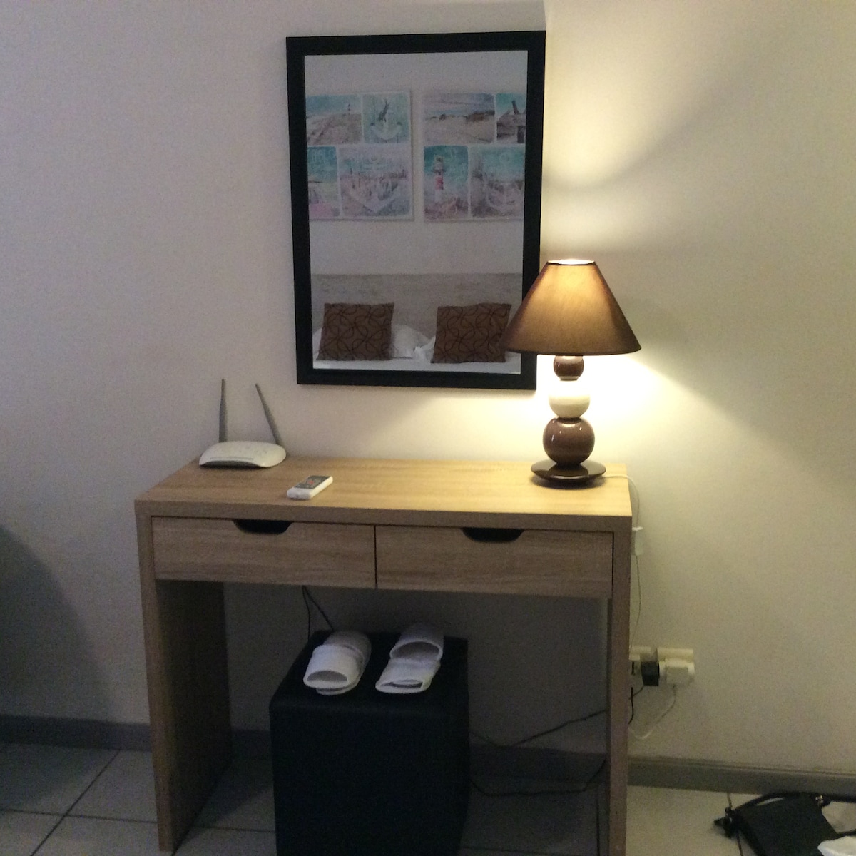 A wooden console table is positioned against a wall. It features two drawers and is accompanied by a table lamp with a warm glow. A mirror hangs above, reflecting the bed area. Slippers are neatly placed on a small black stool beside the table.