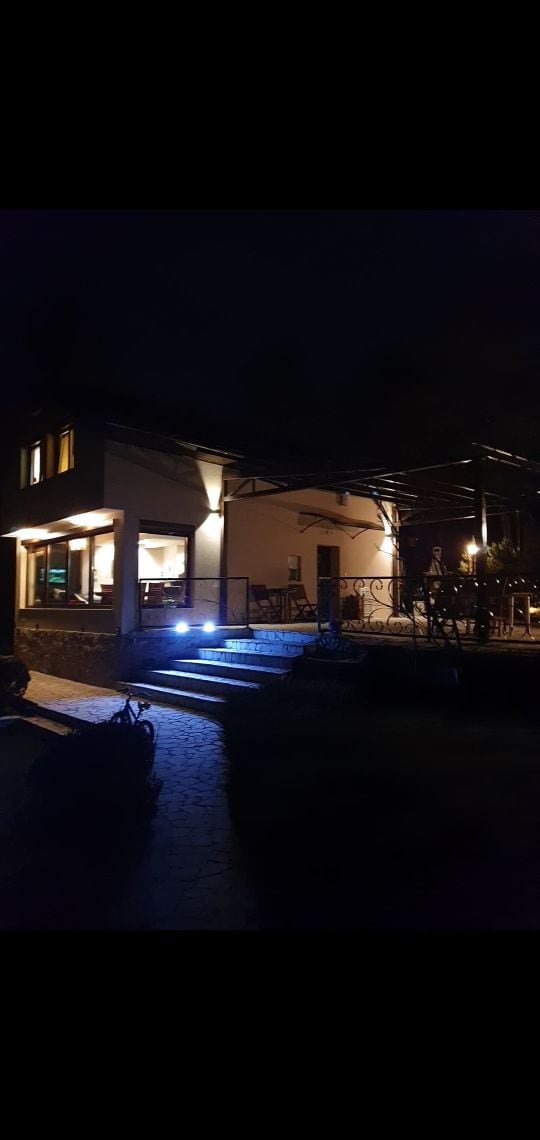 A multi-level villa is shown at night, featuring warm light emanating from windows. A paved walkway leads to a covered terrace, illuminated by soft lighting. The surrounding area is calm and serene, showcasing the house's inviting entrance.