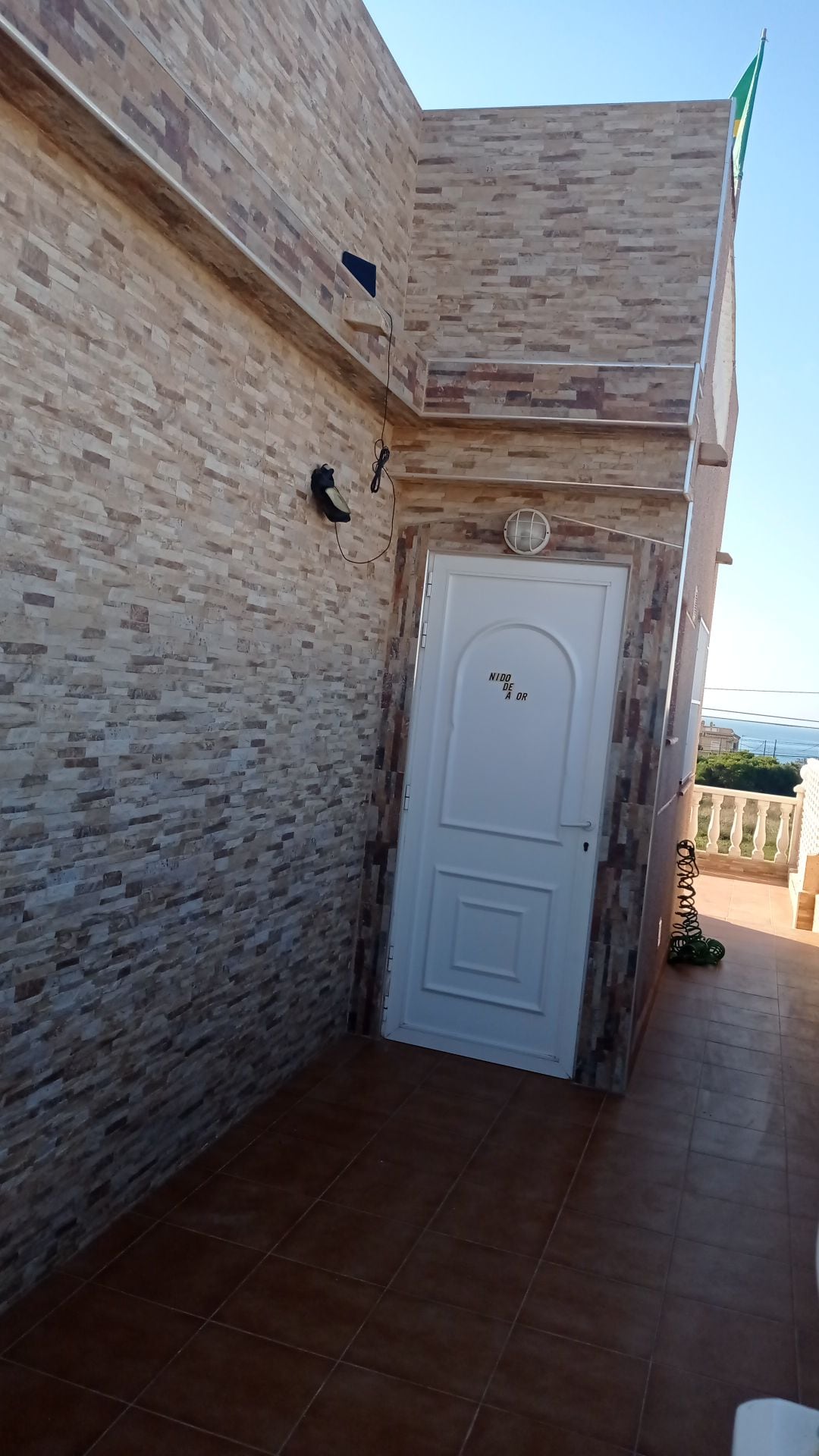 The exterior entrance features a white door with a round clock affixed above it, surrounded by textured stone walls. The tiled floor leads to a spacious area with a view of the surrounding landscape.