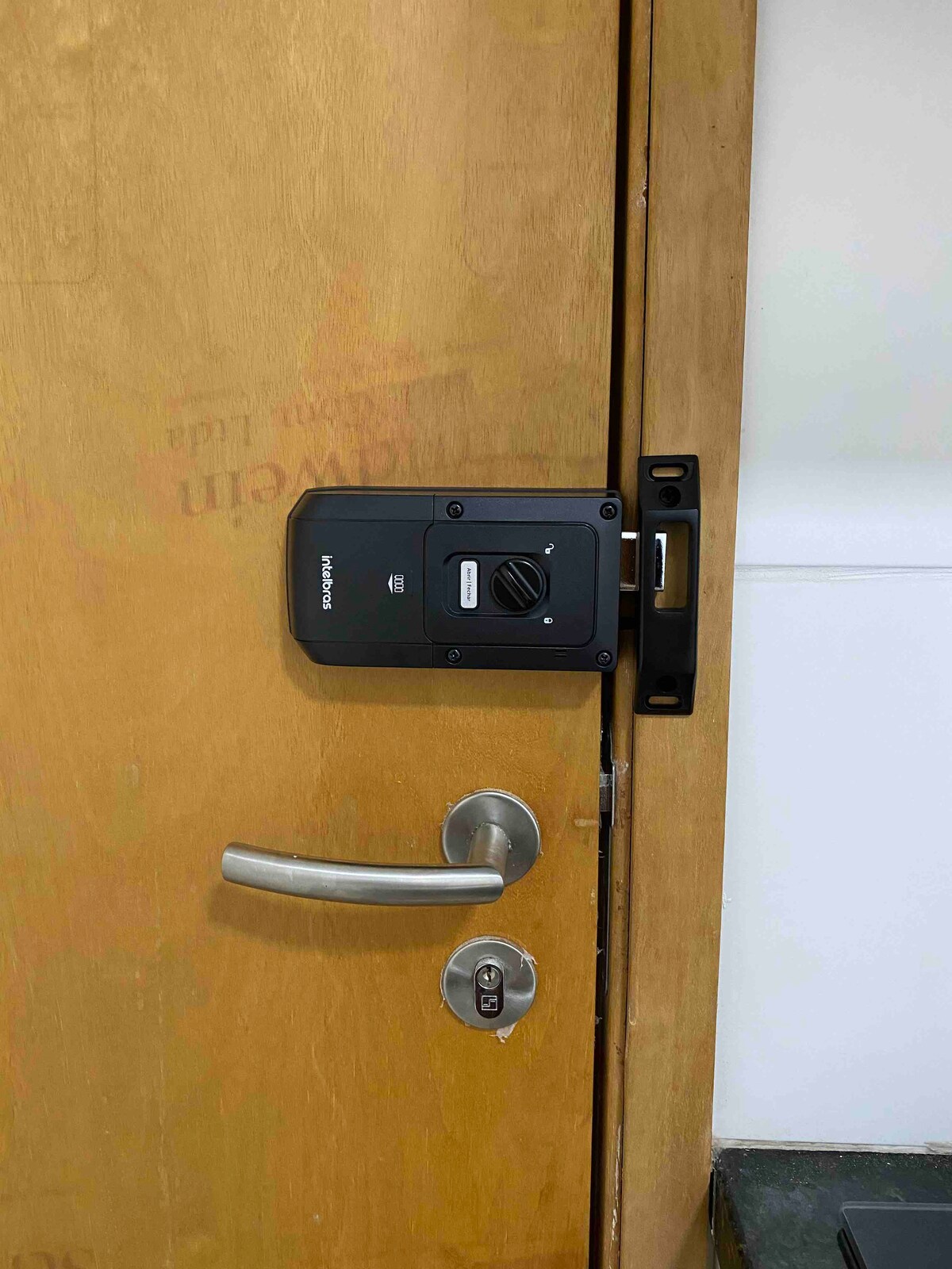 The image displays a sturdy wooden door equipped with a modern locking mechanism. The handle is metallic, and a keyhole is visible below the handle. The door shows signs of use, with some markings on the wood.