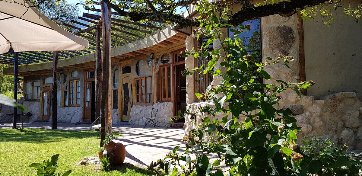 A bioconstructed structure is surrounded by lush green grass and native plants. Natural materials such as stone and wood are visible in the architecture. Sunlight filters through a canopy of leaves, casting gentle shadows on the outdoor seating area, enhancing the connection with nature.