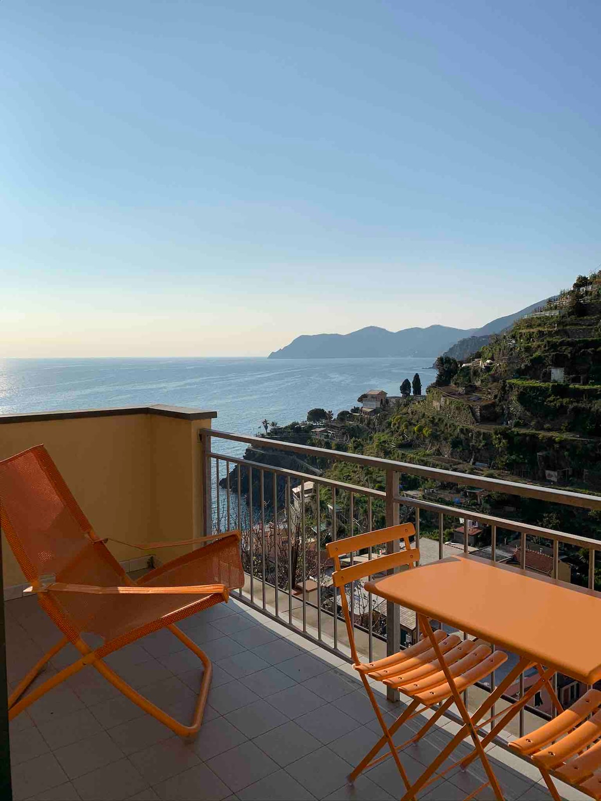 A terrace offers a stunning ocean view, featuring vibrant orange chairs and a matching table. The peaceful scenery includes distant hills and a clear sky, creating a serene outdoor space ideal for relaxation.