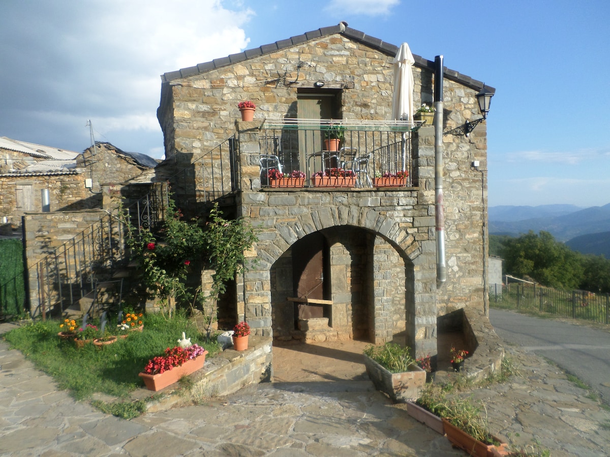 A charming stone building with a rustic façade is framed by vibrant potted flowers. An archway leads into the entrance, while a balcony is adorned with chairs and a table, offering expansive views of the surrounding landscape.