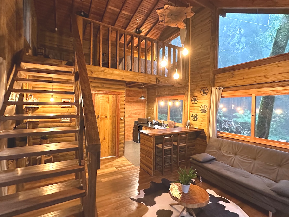 An open interior features a wood staircase leading to a lofted area. Natural light filters through large windows, illuminating a cozy lounge with a comfortable couch and a wooden bar area. Warm wood tones create a harmonious, rustic ambiance.