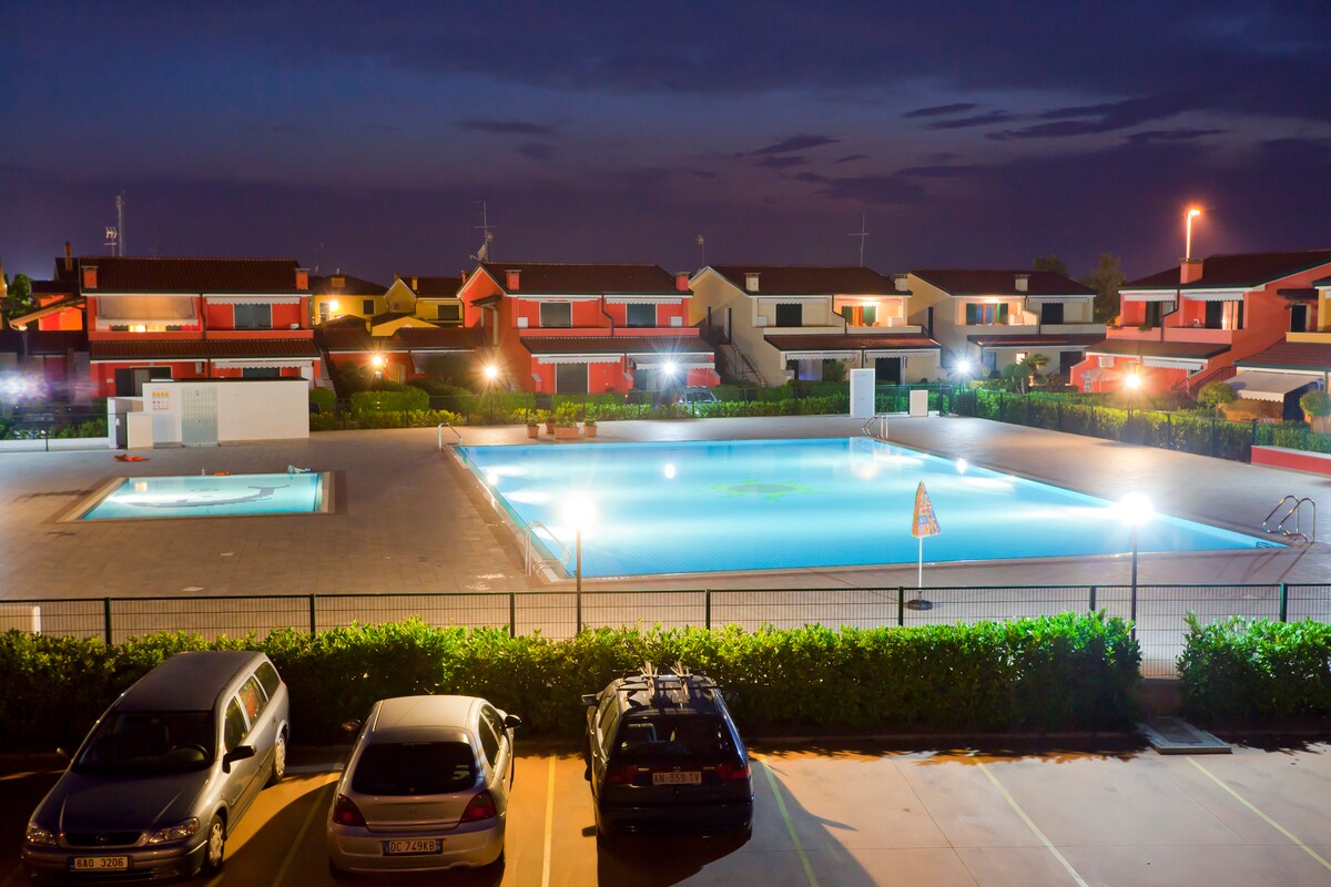 An evening view of the resort features two swimming pools, one larger and one smaller. Surrounding greenery is illuminated, enhancing the tranquil atmosphere. Nearby, parked vehicles are visible, and several accommodations can be seen in the background, with subtle lighting reflecting from their facades.