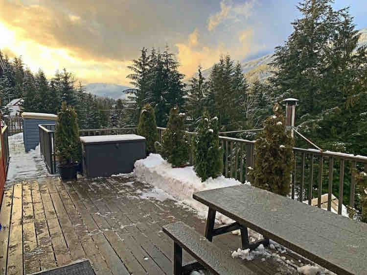 A spacious outdoor deck is surrounded by evergreen trees, with a picnic table positioned for dining. A hot tub is visible, complemented by snow-covered areas and a scenic view of the sky at sunset, creating a serene outdoor space.