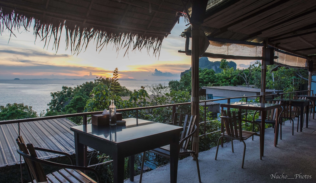 An outdoor dining area features several tables and wooden chairs positioned on a terrace overlooking the ocean. Lush greenery surrounds the space, and a sunset casts warm colors across the sky, highlighting the tranquil atmosphere.