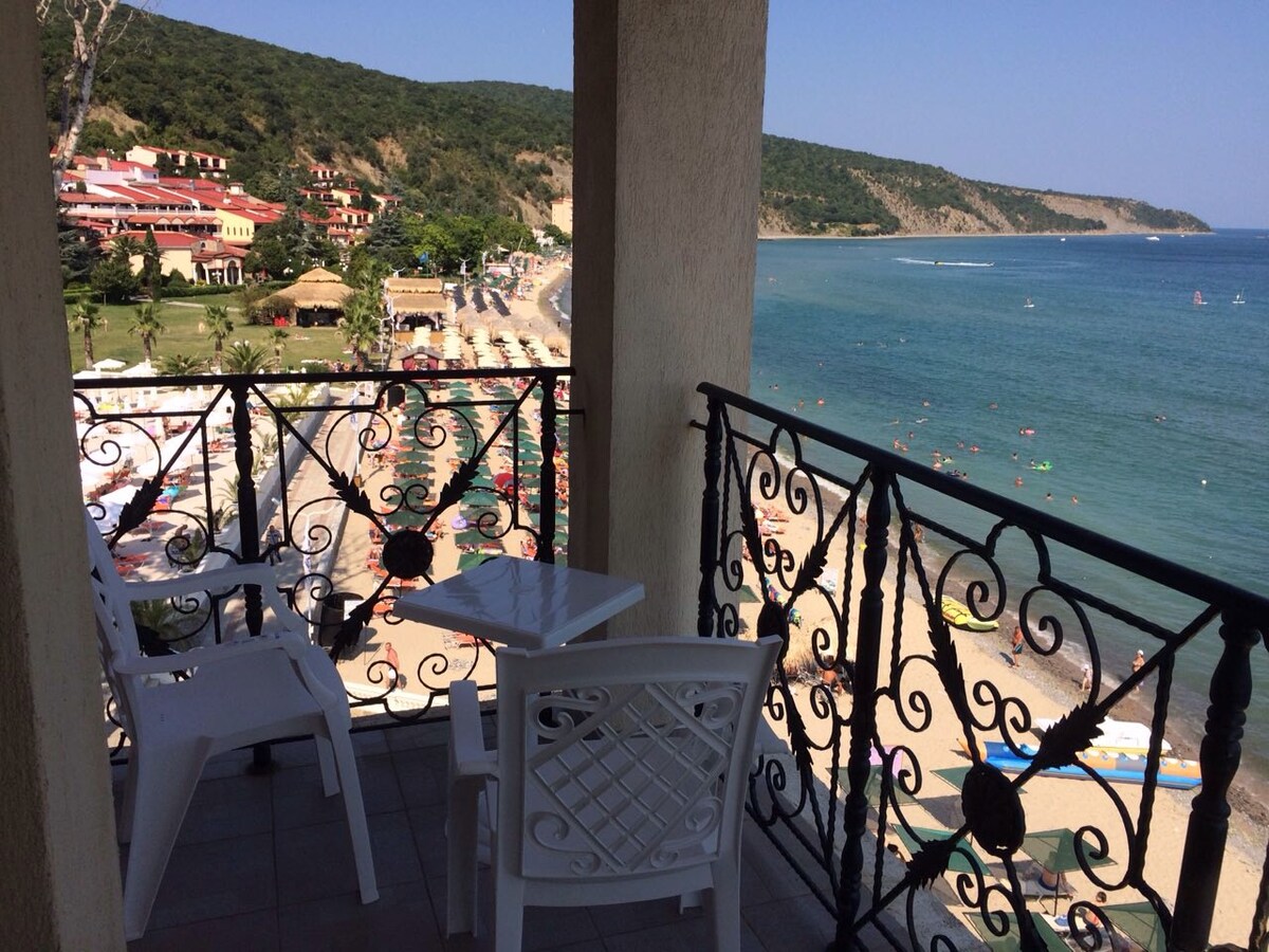 A balcony with ornate black railing overlooks the beach and sea. Two white plastic chairs and a small table are visible, providing a space to enjoy views of the sandy shore and lush green hills in the background under clear blue skies.