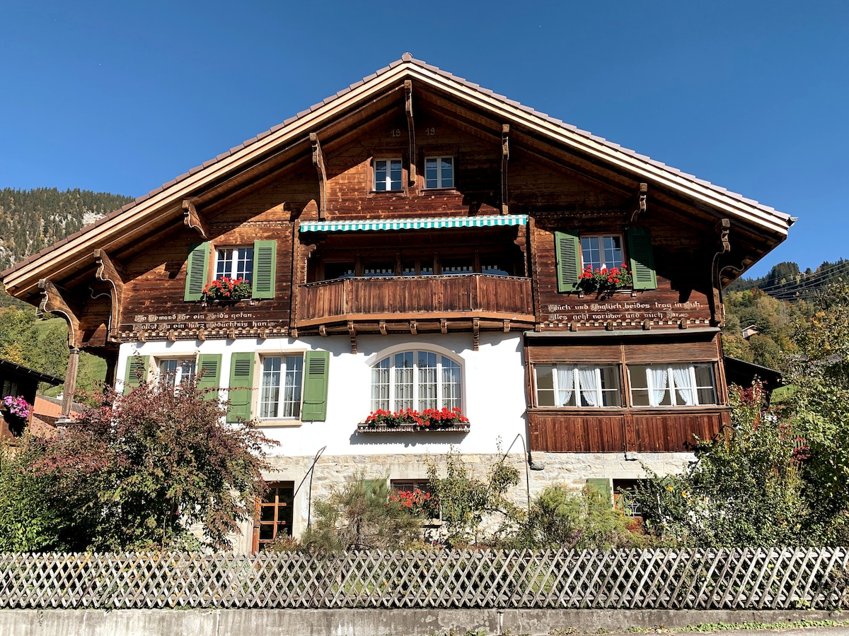 A traditional chalet features a charming wooden facade with decorative balconies adorned with vibrant flower boxes. The structure is complemented by green shutters and a clear blue sky, enhancing its rustic appeal. The surrounding greenery and garden area contribute to the inviting atmosphere.
