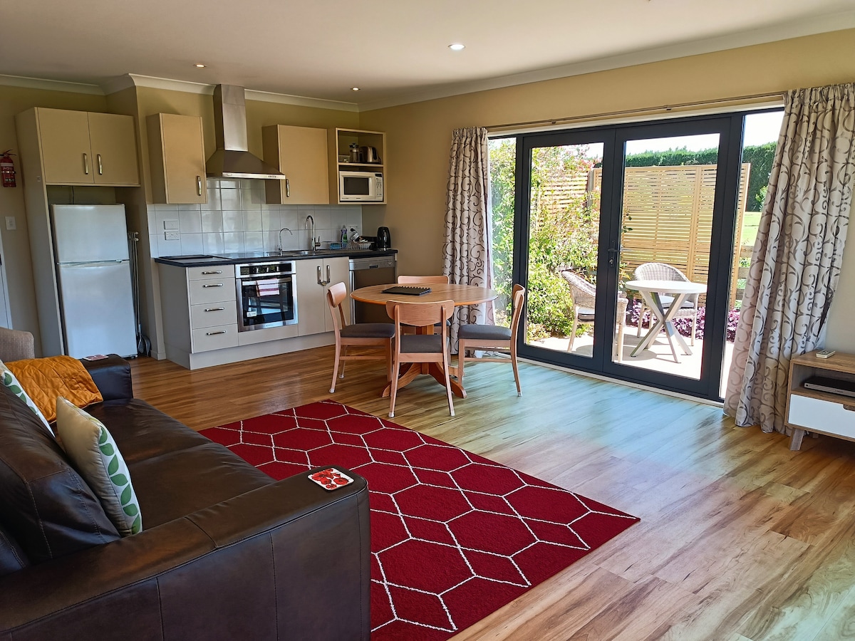 An open plan living area showcases a modern kitchenette equipped with appliances and a dining table for four. Natural light flows through sliding glass doors, leading to a private outdoor seating area. A comfortable sofa and decorative rug enhance the inviting space.
