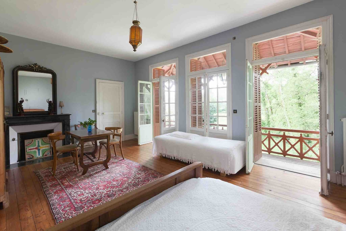 A spacious room features large windows with natural light streaming through. A wooden floor complements a traditional area rug, while a table with chairs invites relaxation. Two open doors lead to a balcony overlooking greenery, enhancing the connection to the outdoors.