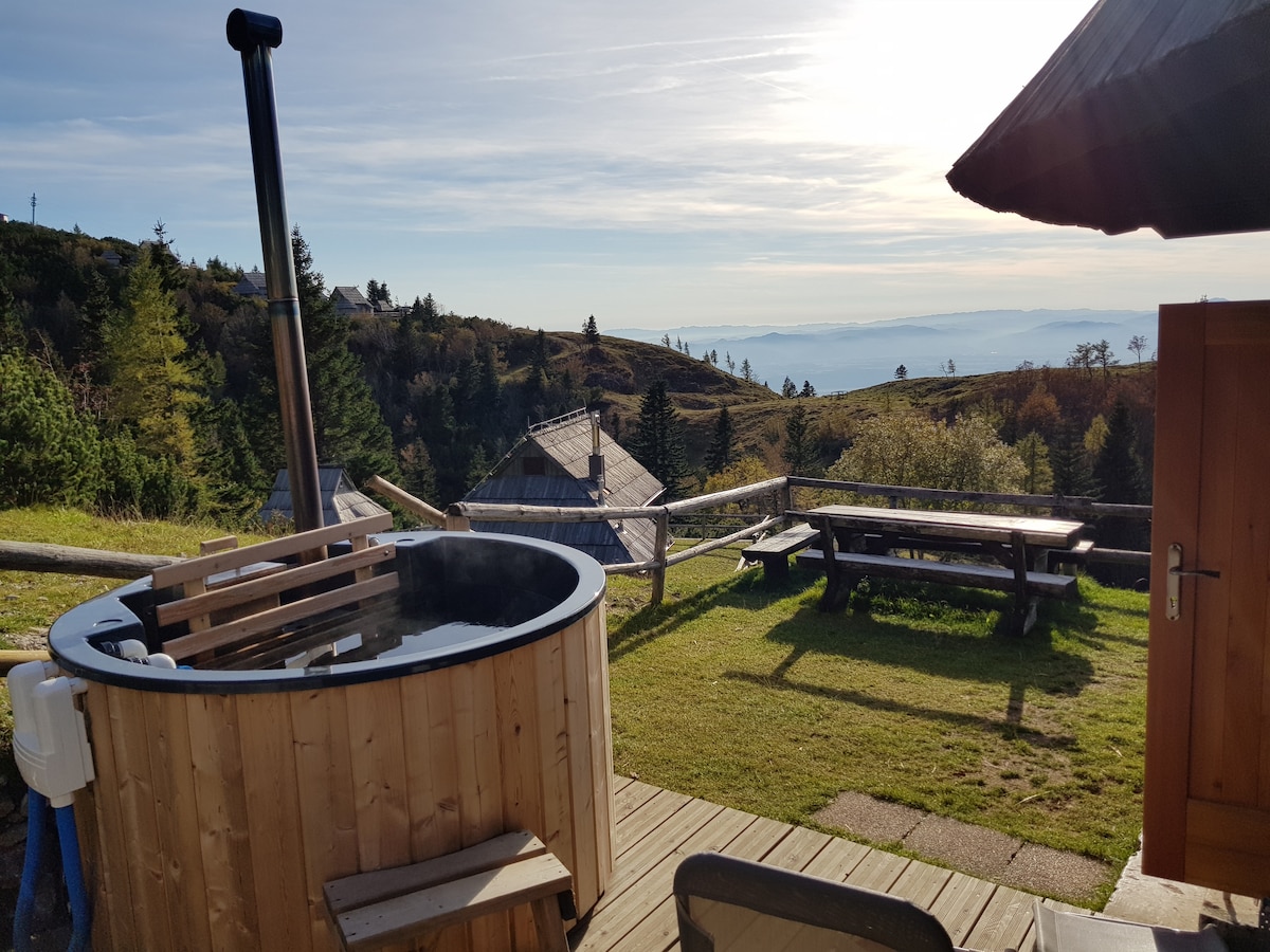An outdoor hot tub made of wood is positioned on a wooden deck, with steam rising from the water. Scenic views of rolling hills and trees extend into the distance, while a nearby picnic table invites outdoor gatherings.