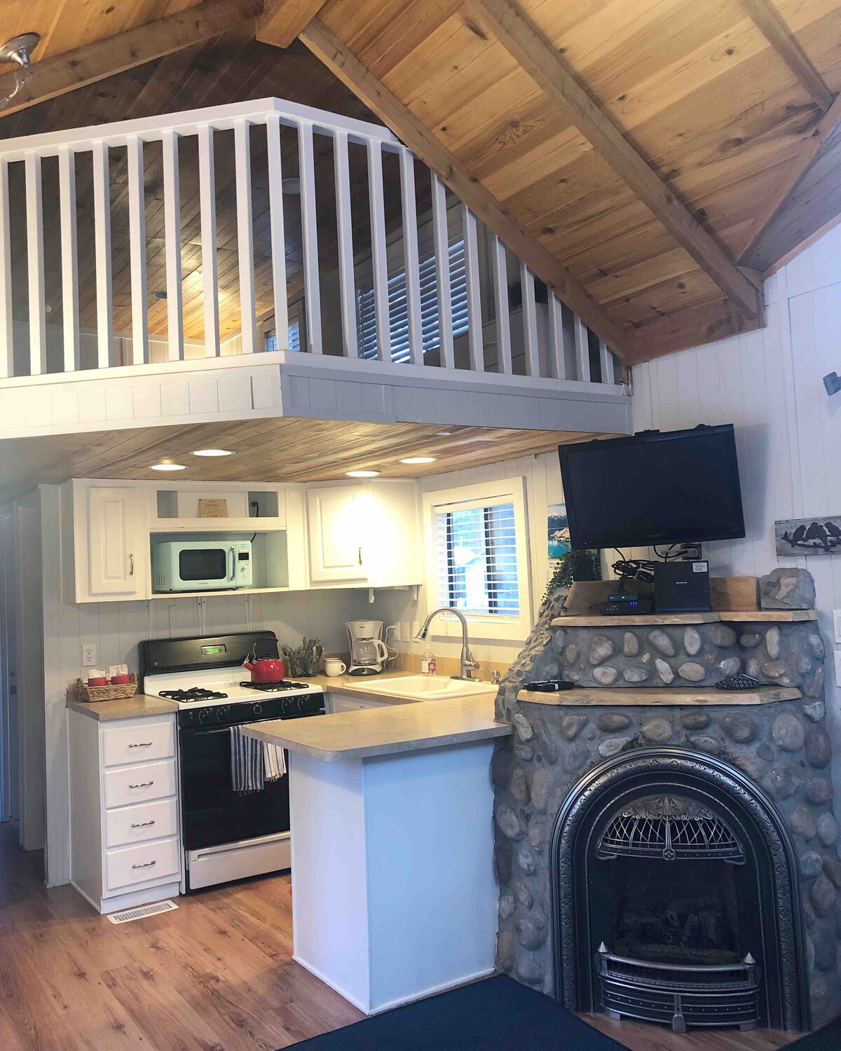 A cozy kitchen area with modern appliances is paired with a warm stone fireplace, creating a functional and inviting space. The lofted area above features a railing with views of the main floor. Natural light filters through the kitchen window, enhancing the cabin's charm.