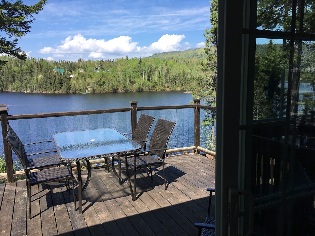 A spacious outdoor deck features a glass-top table surrounded by four chairs, overlooking a calm lake. Lush green trees line the water's edge, and a blue sky with scattered clouds enhances the tranquil setting.