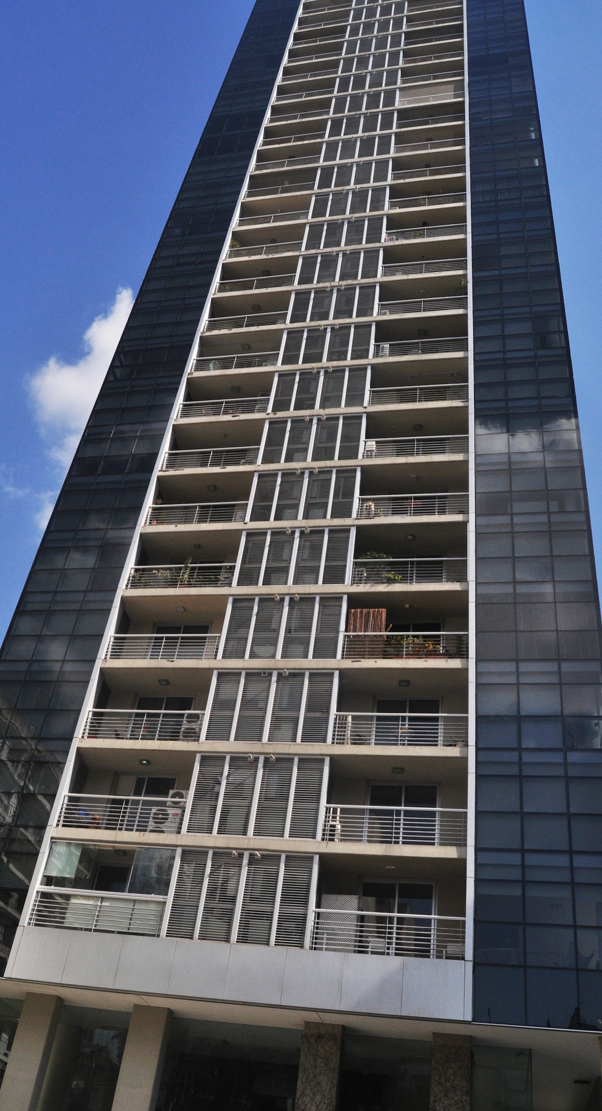 A tall residential building is depicted, showcasing a modern glass facade. Balconies with railings are visible on several levels, providing space for plants or furniture. The structure is set against a clear blue sky, emphasizing its height and contemporary design.