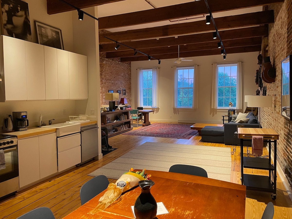 The open living space showcases high ceilings with exposed beams and large windows allowing natural light. A dining table with seating for six is visible, along with a well-equipped kitchen. A comfortable seating area features a dark sofa and colorful area rugs on a polished wooden floor.