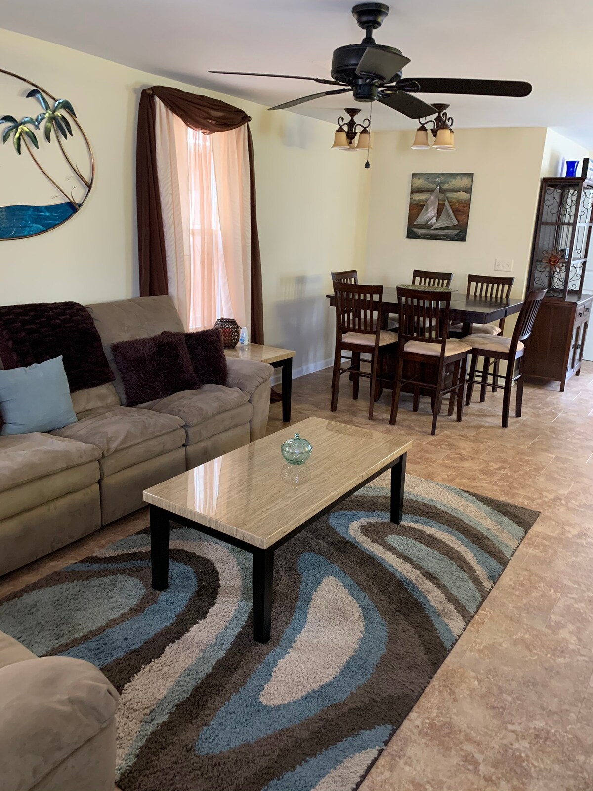 A living area features a comfortable beige sofa with decorative pillows, complemented by a patterned area rug. A glass coffee table stands in the center, and a dining table with chairs is visible in the background. Light filters through the window, creating a relaxed environment.