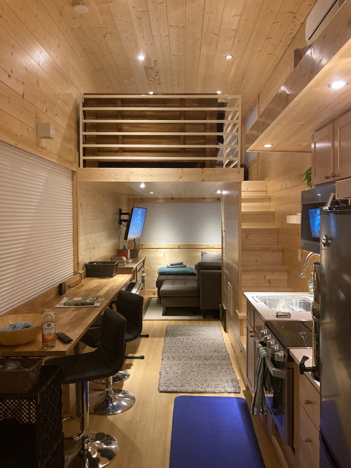 The interior of the tiny house features wooden paneling and a lofted sleeping area visible above. A seating area is positioned beside a compact kitchenette, which includes modern appliances. Bar stools are placed along a narrow counter, enhancing the functional space.