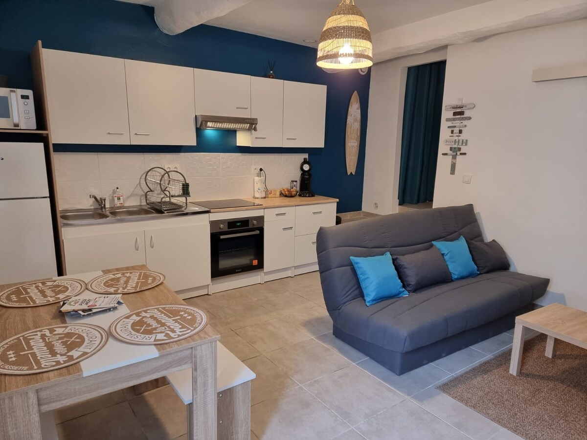A modern kitchen area with white cabinetry and a dark countertop is visible. A gray convertible sofa with teal cushions is positioned in front of the kitchen. A dining table with four chairs and decorative placemats completes the space. Soft lighting is provided by a woven pendant lamp.