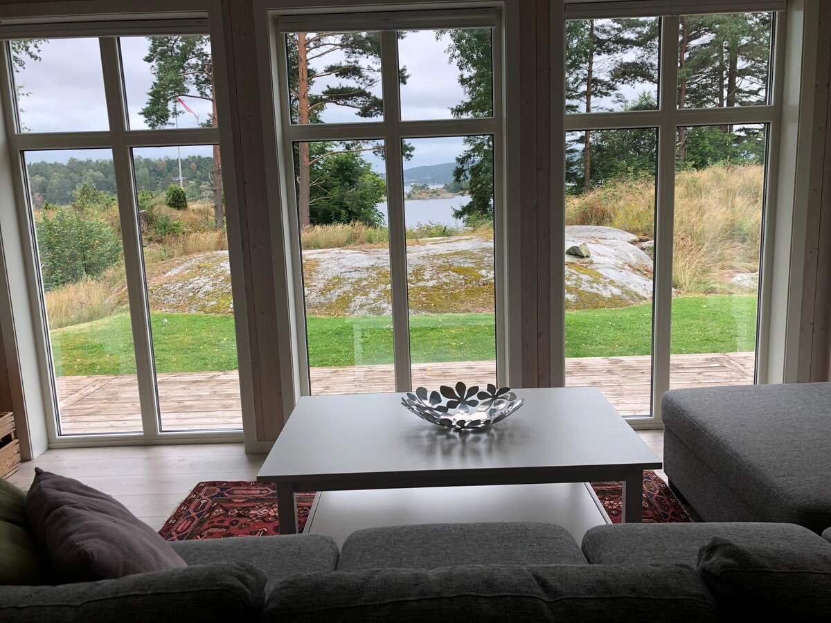 Large windows allow natural light to fill the living space, providing a view of a serene outdoor landscape with grass and rocks. A simple coffee table, adorned with a decorative bowl, is positioned in front of a plush sectional sofa.