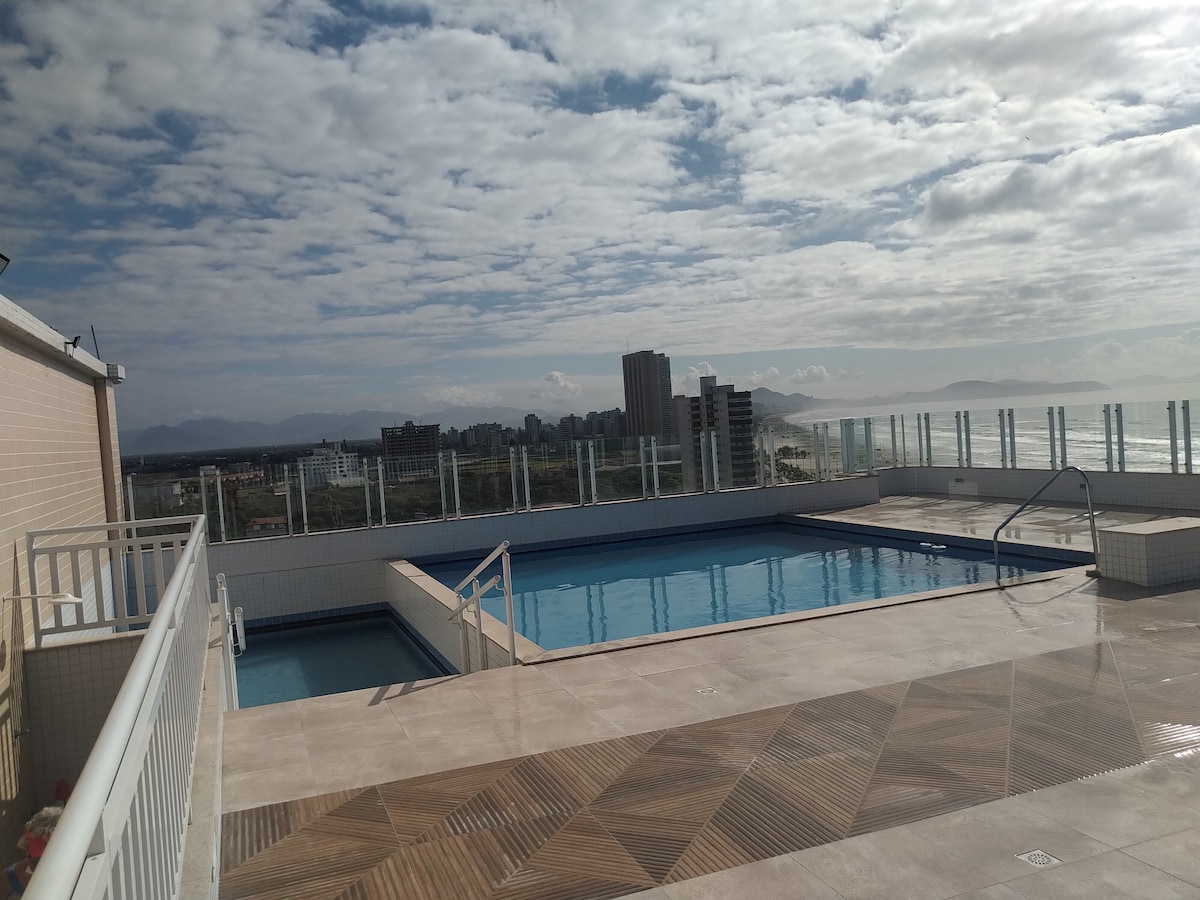 An outdoor pool area is presented with two swimming pools, one larger than the other, surrounded by a spacious sun deck. A panoramic view of the city skyline and distant mountains is visible under a partly cloudy sky.