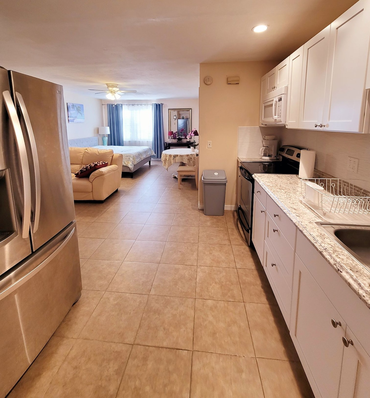 A spacious studio layout showcases a modern kitchen with stainless steel appliances, including a refrigerator. Natural light fills the room, highlighting a cozy seating area with a light-colored couch and a dining table. A comfortable bed is visible in the background, offering convenience and accessibility.