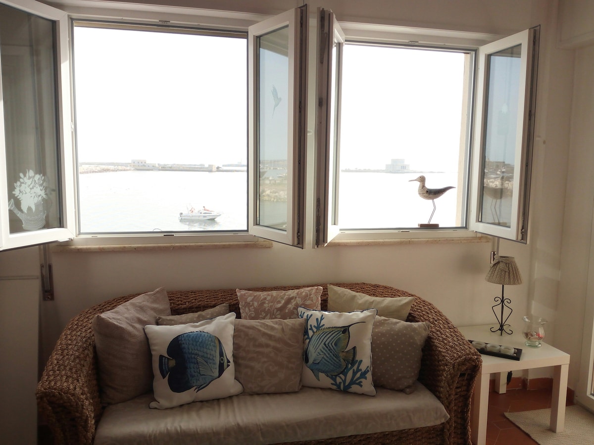 A cozy seating area features a woven sofa adorned with decorative cushions, positioned near large windows that frame a view of the sea. Natural light floods the space, highlighting a small side table and a decorative figure by the window.