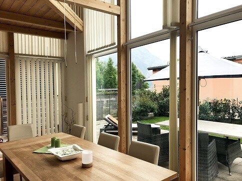 A bright dining area features large windows allowing natural light to fill the space. A wooden table is surrounded by light-colored chairs, with a centerpiece displayed. Outside, a private terrace, green spaces, and comfortable seating can be seen, enhancing the connection between indoor and outdoor environments.