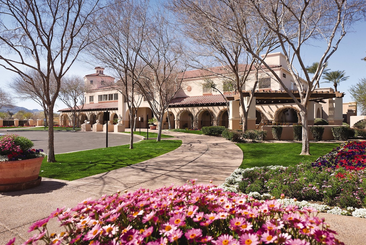 A welcoming resort entrance is framed by blooming flower beds, featuring vibrant pink and white blooms. A winding pathway leads toward a large, adobe-style building with arched doorways and a terracotta roof, surrounded by manicured grass and leafless trees.