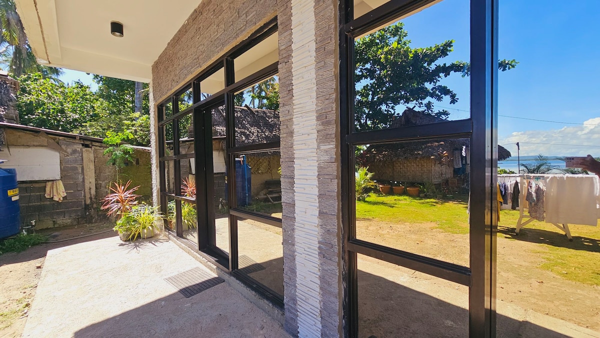 A modern exterior features large glass windows, reflecting the lush greenery and the sky. The view extends to a well-kept garden area, with laundry hanging in the background and a glimpse of outdoor storage and palm trees enhancing the space.