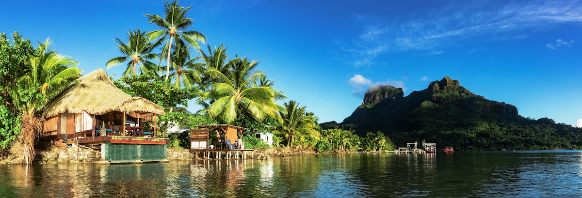 A serene view of a bungalow positioned over the tranquil lagoon, surrounded by lush palm trees. The mountain backdrop provides a stunning contrast against the clear blue sky. Reflections in the calm water enhance the natural beauty of the scene.