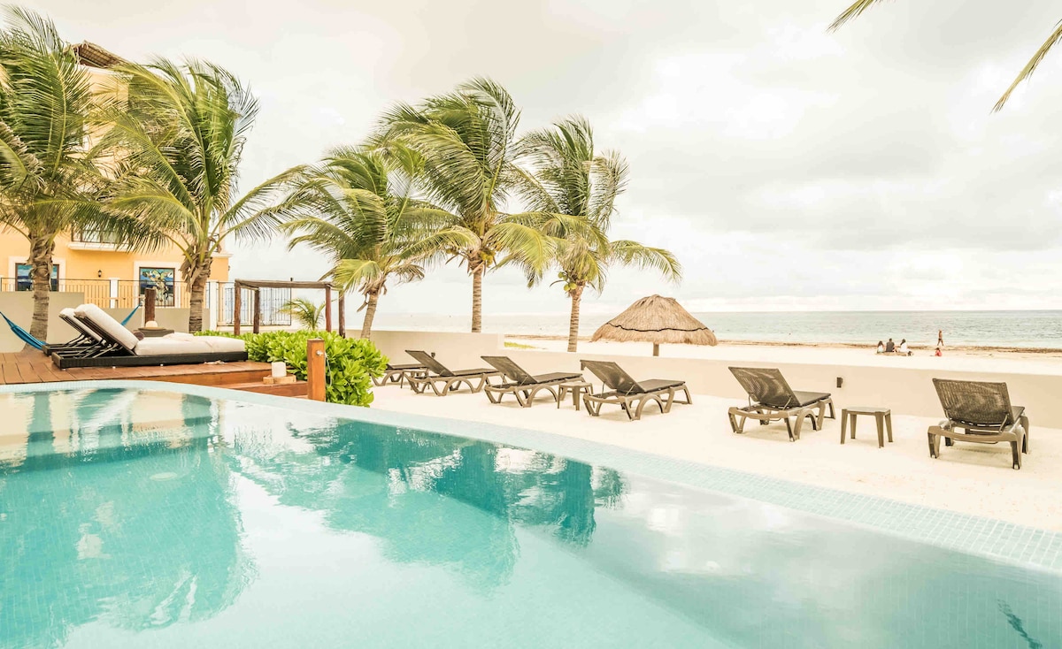 An inviting scene features a clear pool reflecting palm trees and a sandy beach. Several lounge chairs are arranged under the palm trees, with a thatched-roof palapa visible nearby. The tranquil ocean stretches in the background, promising a serene atmosphere.