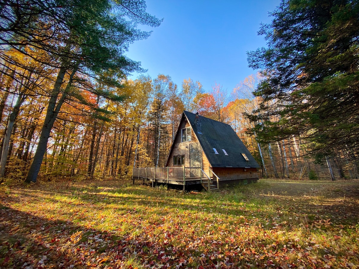 An A-frame cabin stands nestled among colorful autumn trees, showcasing its distinctive peaked roof. A wide wooden deck wraps around the cabin, providing an inviting outdoor space. Fallen leaves cover the ground, creating a natural, seasonal carpet that complements the woodland setting.