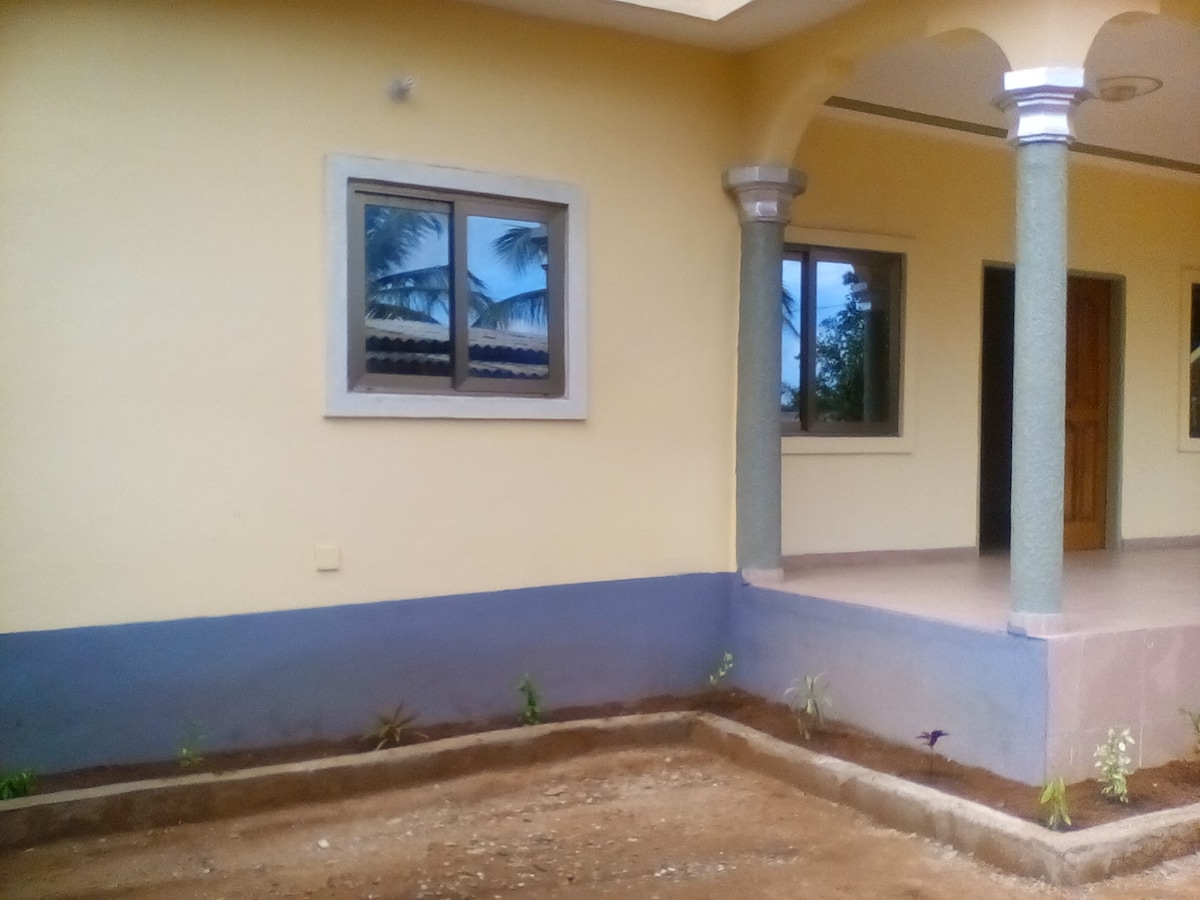The exterior of the villa displays a smooth yellow facade with blue accents at the base. A well-maintained garden area features sparse landscaping. Two windows, framed in gray, allow natural light to enter, complemented by structural columns providing architectural detail.