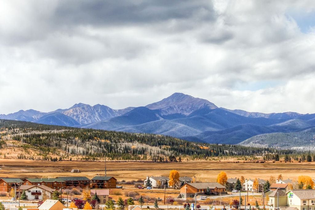 A panoramic view showcases the majestic mountains, framed by a landscape of rolling hills and dotted with trees. The seasonal colors reveal golden foliage alongside patches of green, while a serene sky provides a dramatic backdrop with soft, cloud-covered tones.
