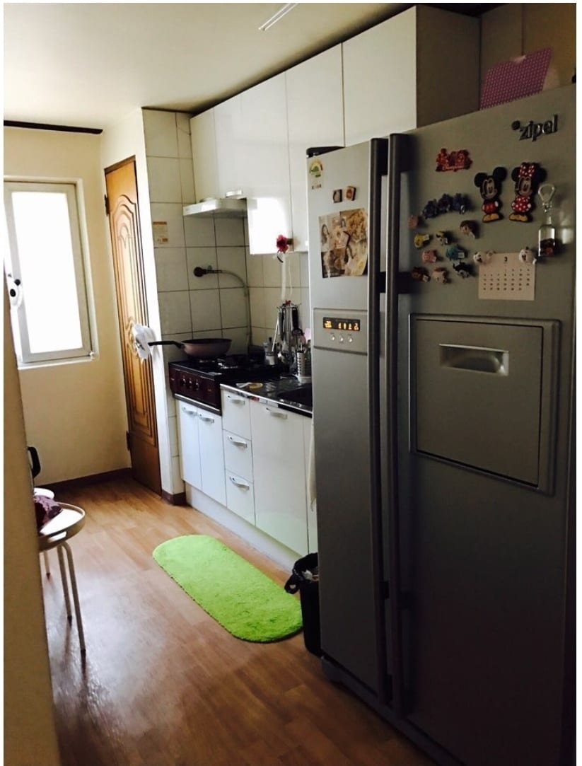 The kitchen features modern white cabinetry and a stainless steel refrigerator. A black stove and sink are positioned against the wall, complemented by a small wooden table with a chair. Natural light enters through a window, illuminating the space.