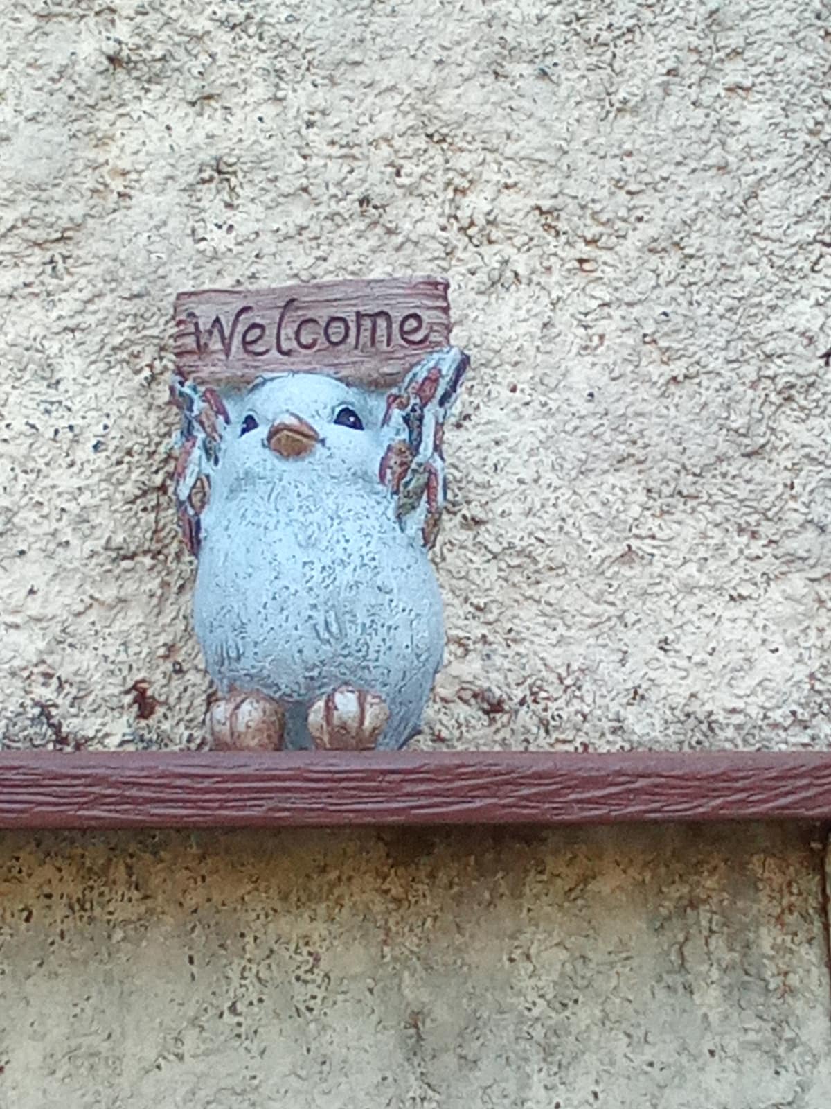 A decorative bird figurine is perched on a shelf, holding a small sign that reads 'Welcome.' The textured surface of the bird's body contrasts with the smoothness of the sign. The background features a rough, sandy wall, adding warmth to the scene.