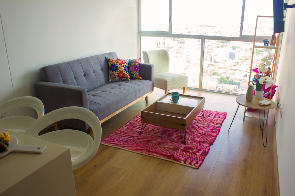 A modern living area features tall windows offering expansive city views. A gray sofa is accented by colorful cushions, while a light chair complements a low wooden coffee table, which is placed on a vibrant area rug. The space is filled with natural light.