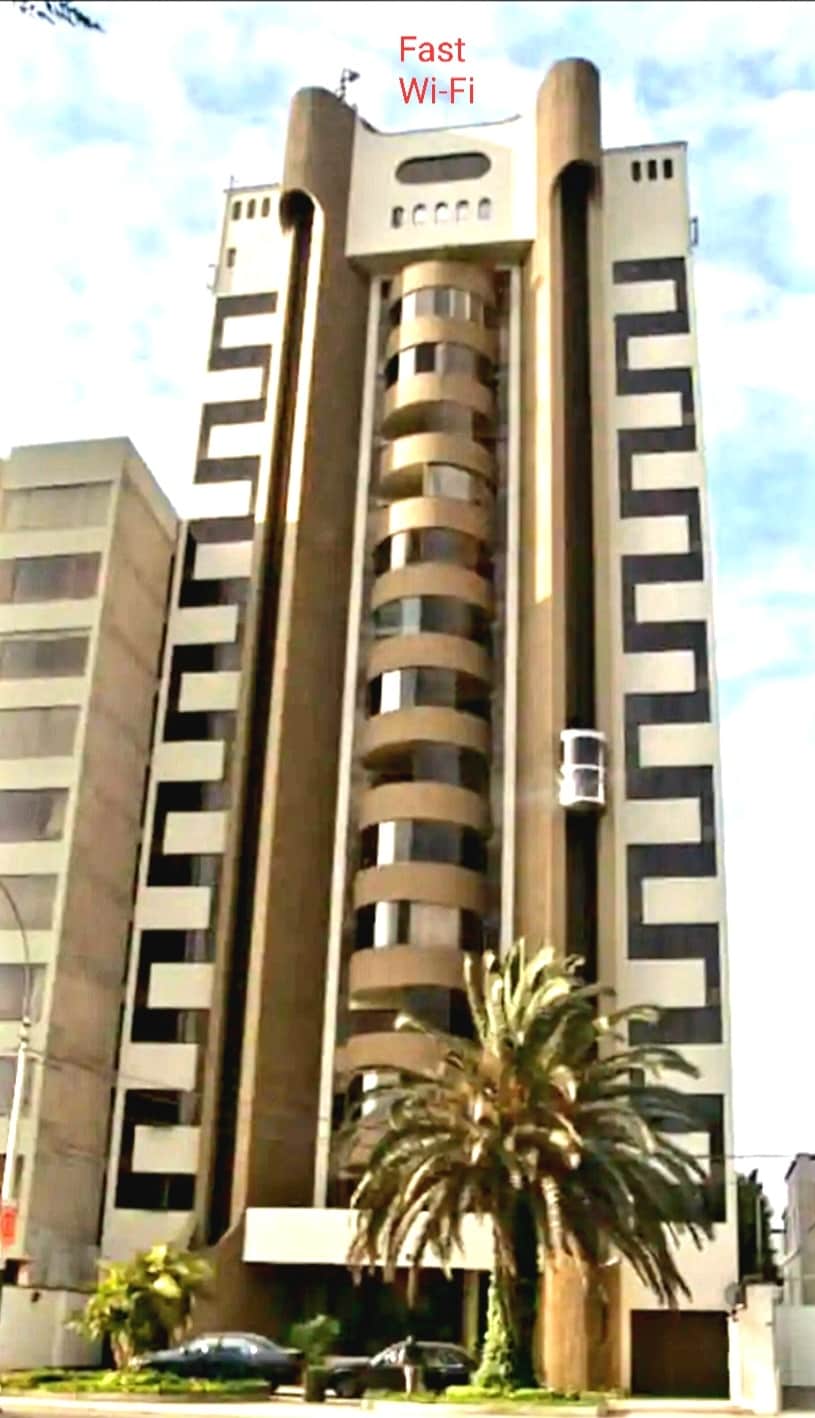 A tall building is captured from the street, featuring a modern architectural design with alternating patterns on the façade. Large windows are visible, complemented by a palm tree at the entrance. A sign indicates fast Wi-Fi availability.