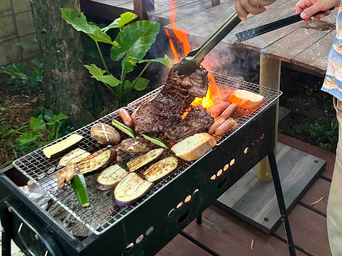 A barbecue grill is seen with flames emerging as various foods are being cooked. Meat, sausages, and slices of vegetables are arranged on the grill, with a hand holding tongs positioned near the food. Lush greenery and wooden decking provide a natural backdrop.