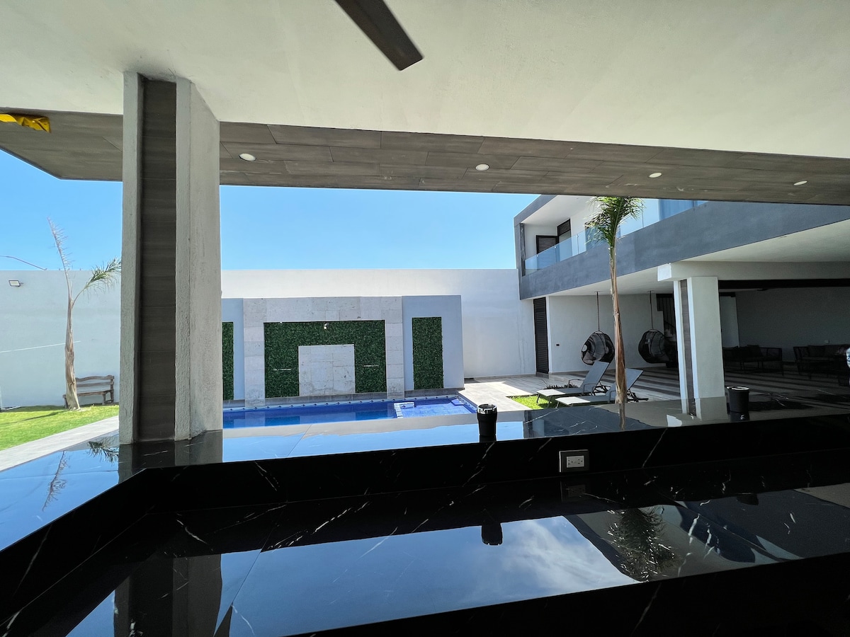 The view from the patio captures a modern exterior featuring a swimming pool surrounded by grass and clean lines. Large sliding doors lead to an inviting living area. Two hammocks are positioned nearby, contributing to a relaxed atmosphere within the open space.