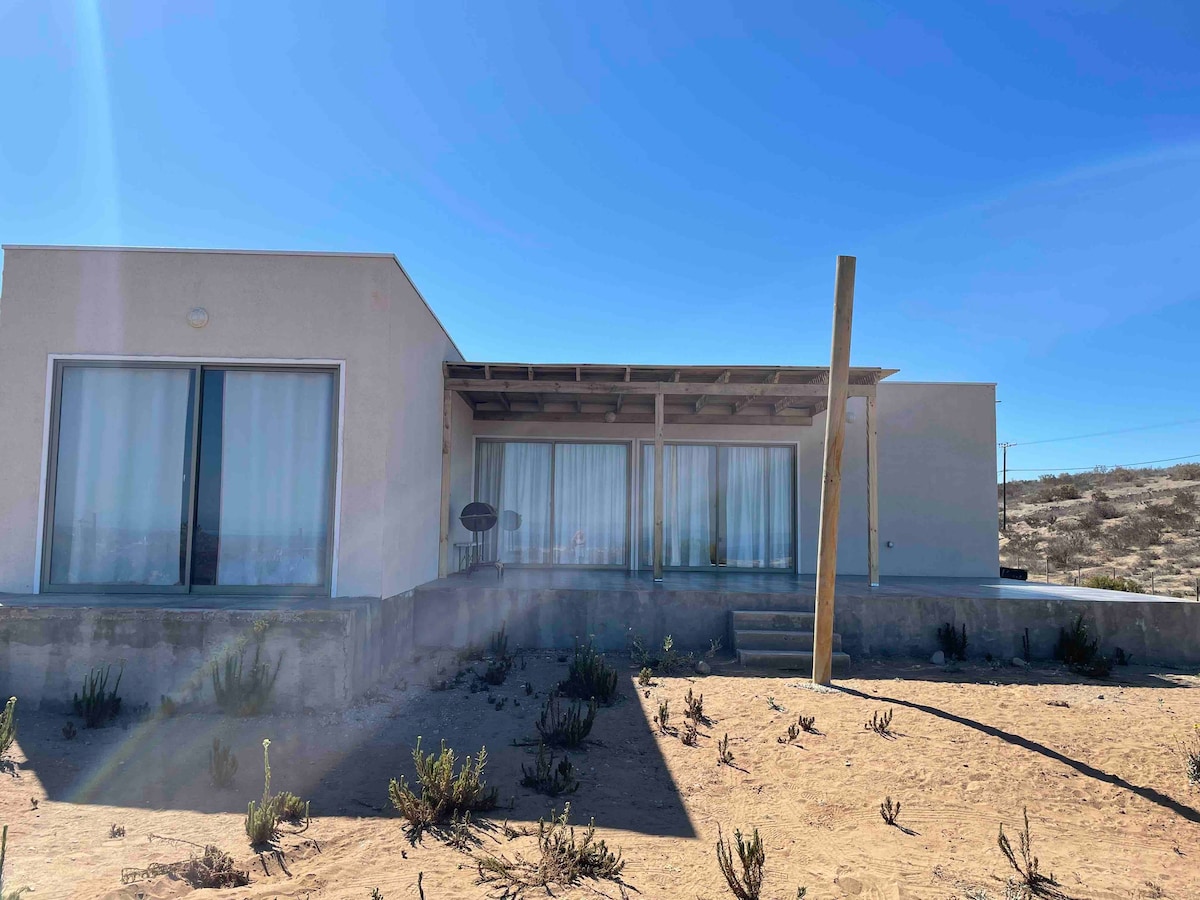 A spacious building is set against a clear blue sky, featuring large glass windows that allow natural light to fill the interior. A wooden support post stands near the entrance, and low vegetation surrounds the property, complementing the desert landscape.
