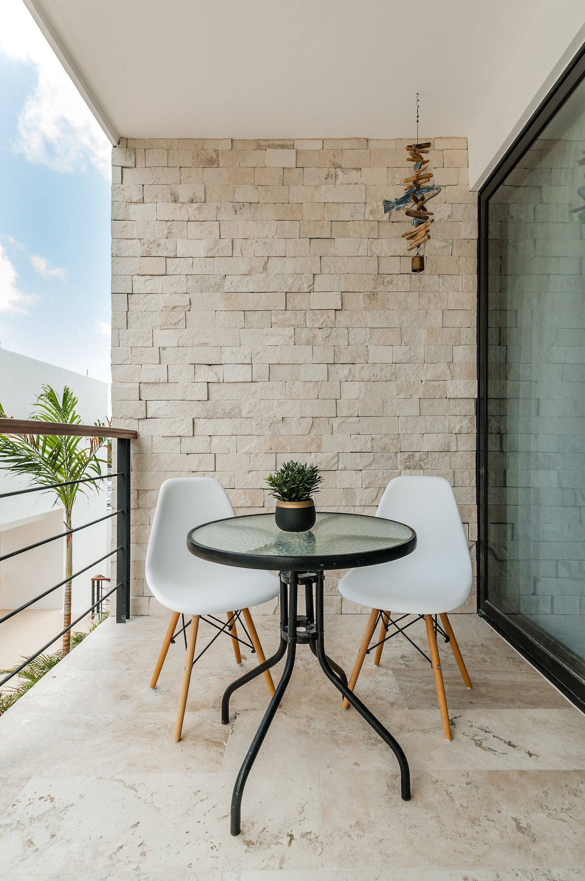 A small balcony features a round glass-top table paired with two white chairs. A decorative plant sits on the table. The backdrop consists of a textured stone wall, complemented by a view of the sky and a nearby palm tree.