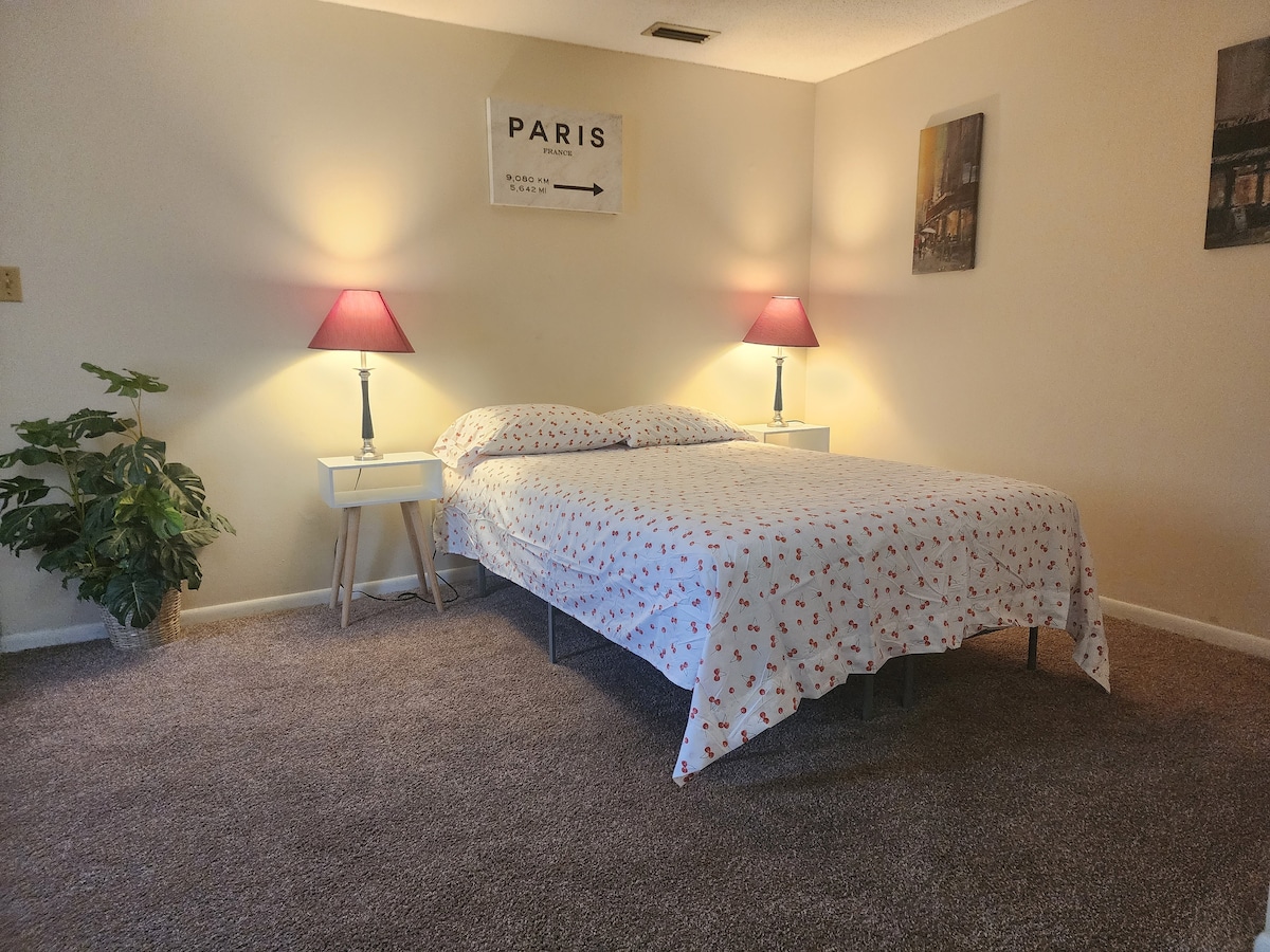A cozy bedroom is furnished with a spacious bed covered in a patterned bedspread. Soft lighting from two bedside lamps creates a warm glow. A framed sign and two wall art pieces enhance the ambiance, while a small green plant adds a touch of nature.