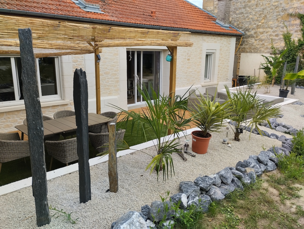 An outdoor terrace is visible, featuring a wooden pergola and a dining table with chairs. Lush potted plants are arranged along pebbled pathways, while decorative stones outline the area. The facade of the stone house can be seen in the background, with large glass doors leading inside.