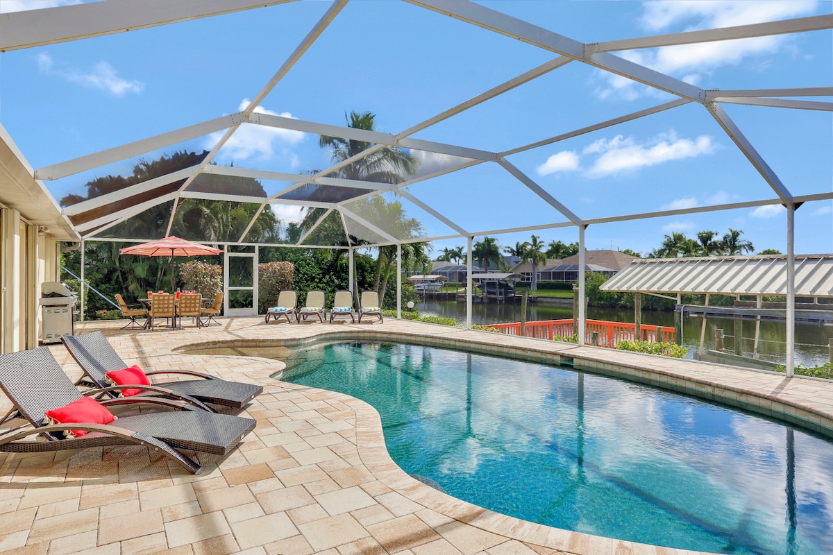 The pool area is surrounded by a screened enclosure, featuring a large freeform pool with a wading area. Multiple lounge chairs are positioned for sunbathing, while a shaded dining area with an umbrella sits nearby. Lush greenery and a waterfront view are visible in the background.
