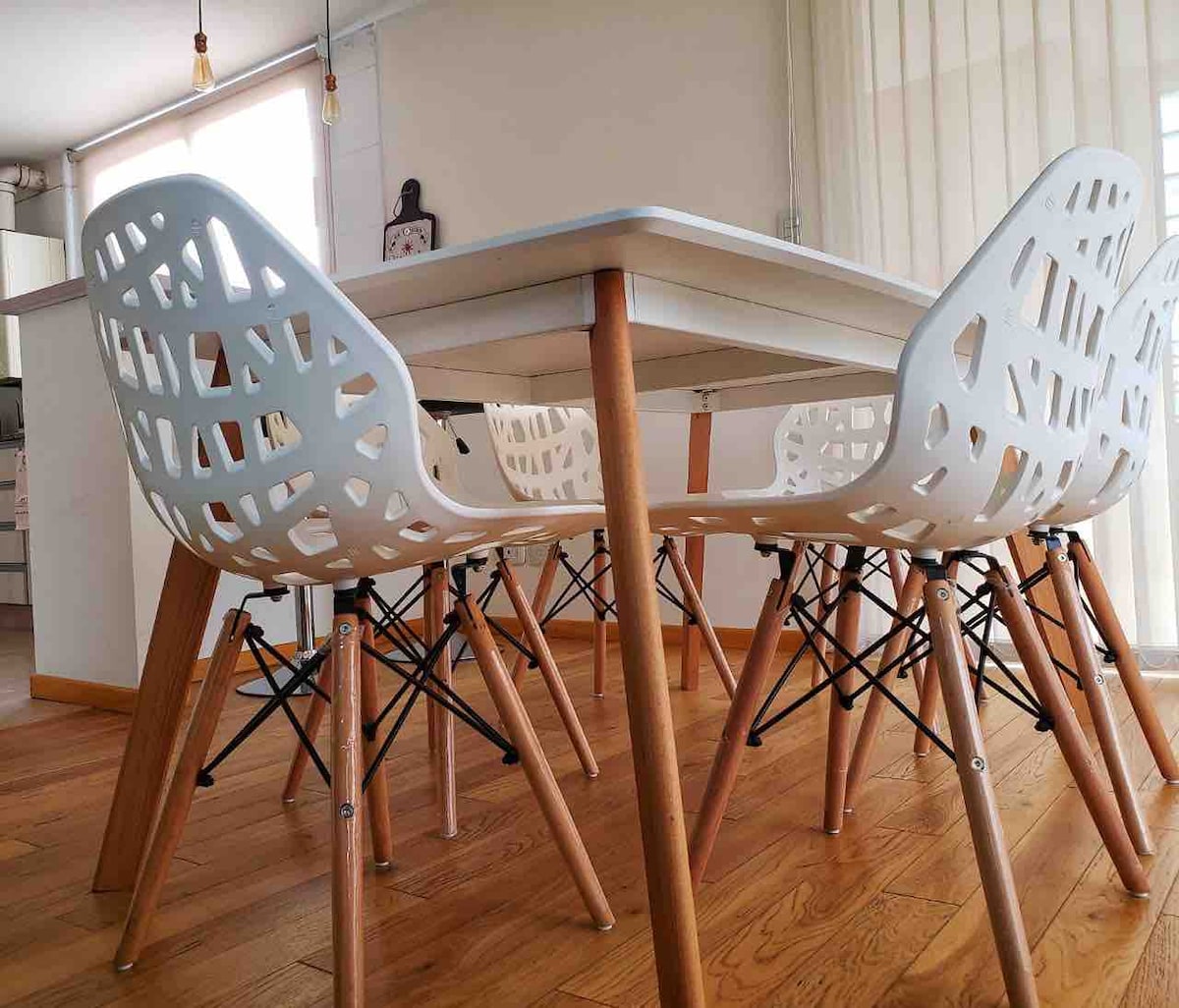A dining table is set with a smooth, white surface and wooden legs. Four contemporary chairs with a unique design surround the table, featuring white plastic seats and slender black metal supports. Natural light filters through sheer curtains, illuminating the warm wooden flooring.