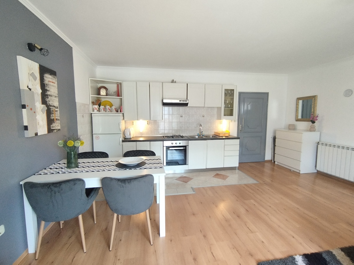 An open kitchen and dining area is presented, featuring a modern layout with white cabinets and appliances. A dining table with two upholstered chairs is positioned beside a bouquet of flowers. Natural light brightens the space, complemented by a light-toned laminate floor.